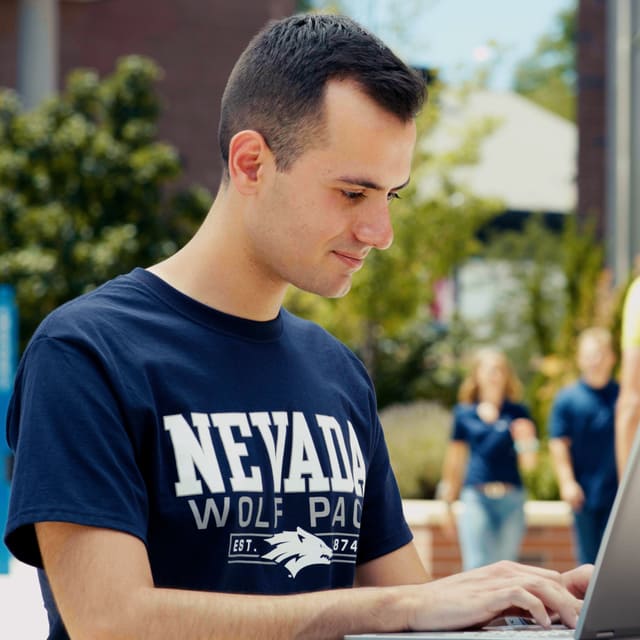 A student wearing a blue Nevada Wolf Pack shirt works on a laptop outside.